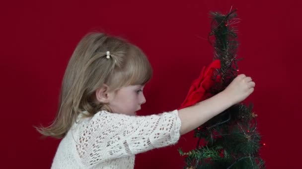 Ein Mädchen in einem eleganten Kleid auf rotem Hintergrund schmückt einen Weihnachtsbaum — Stockvideo