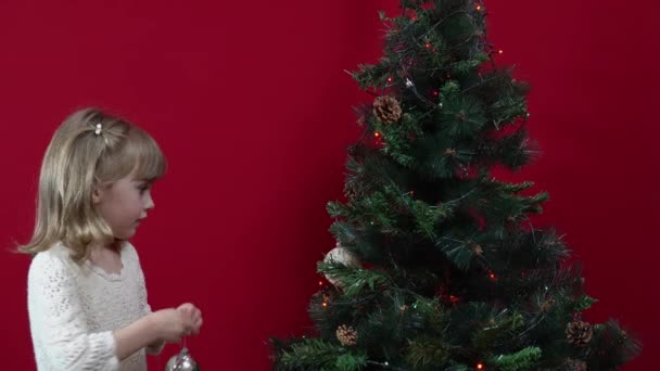 Una ragazza in un abito elegante su uno sfondo rosso decora un albero di Natale — Video Stock