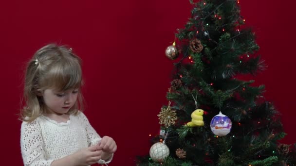 Uma menina em um vestido elegante em um fundo vermelho decora uma árvore de Natal — Vídeo de Stock