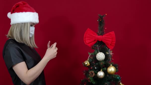 Žena v Santa Claus klobouk a lékařská maska na červeném pozadí fotografování vánoční strom — Stock video