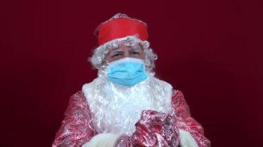 Rus Noel Baba, kırmızı arka planda tıbbi bir maske takıyor ve ışıltılı bir el çırpıcı fırlatıyor.