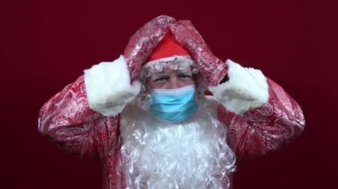 Rus Noel Baba, kırmızı arka planda tıbbi bir maske takmış, üzgün, başını iki eliyle tutuyor.