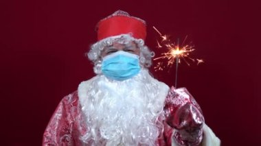 Kırmızı arka planda tıbbi maskeli neşeli Rus Noel Baba elinde bir Binshgal ateşi tutuyor.