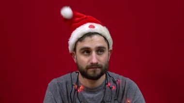 Adam oturuyor ve kafasında bir Noel şapkası dans ediyor. Kırmızı arka planda Noel Baba sakallı yardımcı