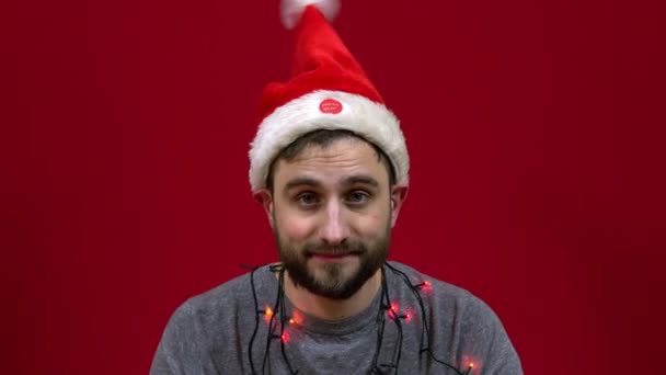 Muž sedí a na hlavě mu tančí vánoční klobouk. Santas vousatý pomocník na červeném pozadí — Stock video