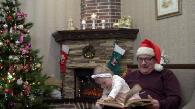 Noel Baba şapkalı büyükbaba torununa diz çöktürdü ve Noel 'den önce şöminenin yanında masal okudu. Yeni yıldan önce neşeli bir atmosfer.