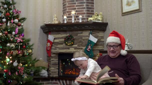 El abuelo con un sombrero de Santa puso a su nieta de rodillas y lee un cuento de hadas junto a la chimenea antes de Navidad. Ambiente festivo antes del nuevo año — Vídeos de Stock