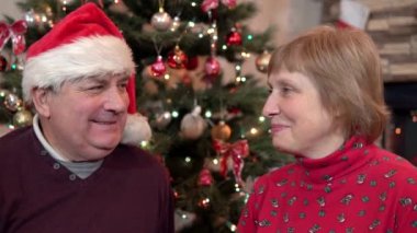 Noel Baba şapkalı bir adam, bir kadına Noel ağacının arka planında Noel hediyesi verir ve sarılır.