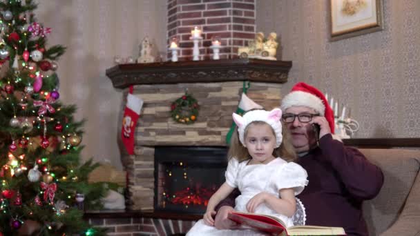 Avô de chapéu de Pai Natal com a neta de joelhos e a falar ao telefone. Atmosfera festiva antes do ano novo — Vídeo de Stock