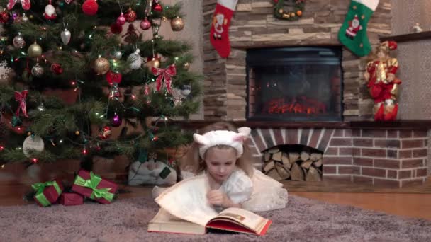 O fată îmbrăcată într-o rochie albă lângă șemineu sub pomul de Crăciun se află pe stomac și citește o carte. Atmosfera festivă înainte de noul an — Videoclip de stoc