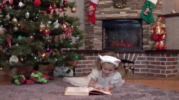 Una ragazza con un vestito bianco vicino al camino sotto l'albero di Natale è sdraiata sul suo stomaco e sta leggendo un libro. Atmosfera festiva prima del nuovo anno — Video Stock