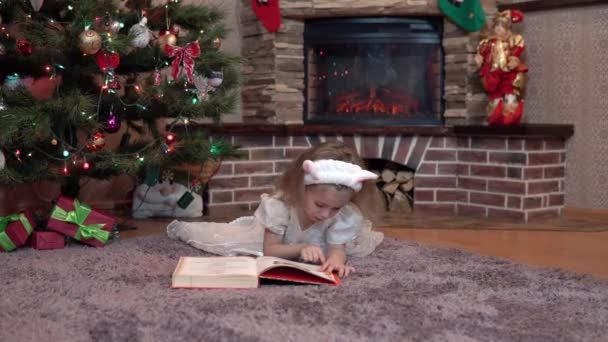 En flicka i vit klänning vid öppna spisen under granen ligger på magen och läser en bok. Festlig atmosfär före det nya året — Stockvideo