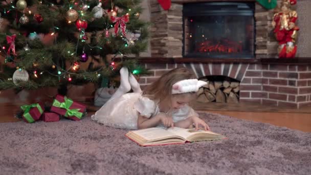 Uma menina em um vestido branco perto da lareira sob a árvore de Natal está deitada em seu estômago e lendo um livro. Atmosfera festiva antes do ano novo — Vídeo de Stock