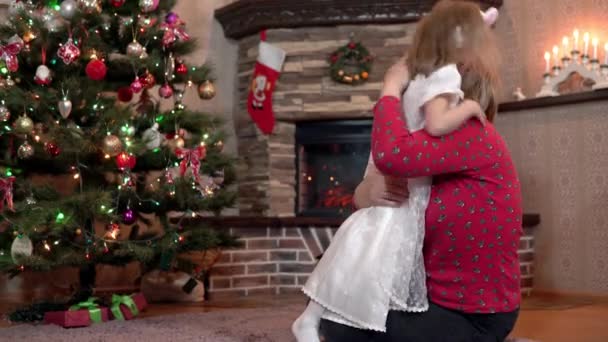 Kleindochter geeft oma een kerstcadeau en kust en knuffelt haar bij de boom — Stockvideo