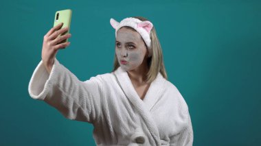 Beyaz cüppeli bir kadın, yüzünde karbonlu balonlu kilden bir maske, 4K selfie çekiyor.