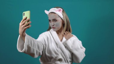 Beyaz cüppeli bir kadın, yüzünde karbonlu balonlu kilden bir maske, 4K selfie çekiyor.