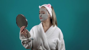 Beyaz cüppeli bir kadın, karbonlu baloncuklu kilden yapılmış bir yüz maskesi aynaya bakıyor.