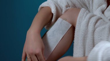 Evinde turkuaz arka planda balmumu epilasyon kullanan genç bir kadın. Vücut Kılları Çıkarma 4K için Balmumu Sıcaklığı