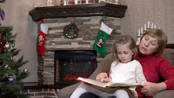 Бабушка посадила внучку на колени и перед Рождеством читает сказку у камина. Праздничная атмосфера перед Новым годом — стоковое видео