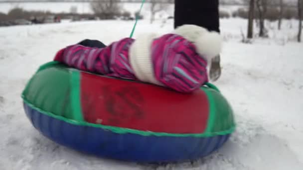 A menina está sendo enrolada em um anel inflável na neve. Uma menina na roupa morna encontra-se em um anel inflável e é puxada por uma corda por uma outra pessoa — Vídeo de Stock