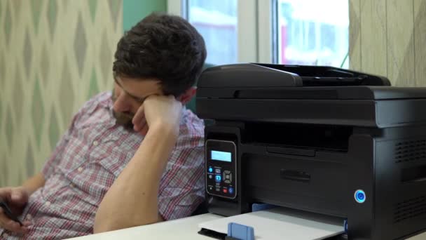 Der Drucker auf dem Schreibtisch druckte dem Mann neben ihm zwei leere Blätter Papier. Mann im Büro nutzt Smartphone am Arbeitsplatz — Stockvideo