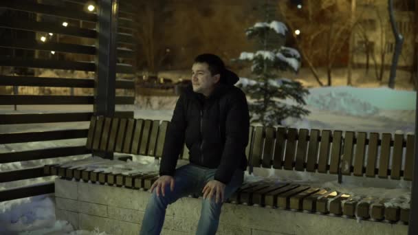 Mężczyzna siedzi na ławce wieczorem w zimie i nie czeka na kogoś niecierpliwie. — Wideo stockowe