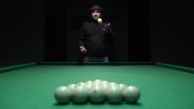 Kapüşonlu bir adam piramide istiflenmiş beyaz topların önüne bilardo topu atıyor.