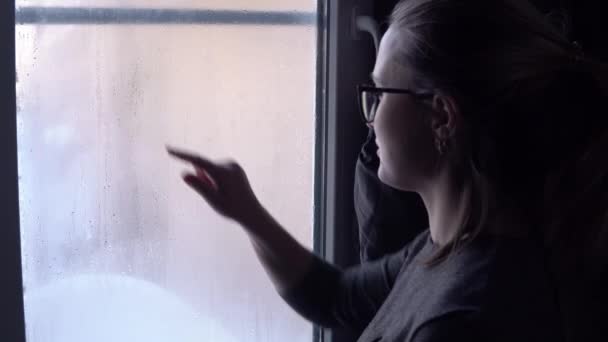 Dívka stojí u zamlženého okna. Píše prstem na zamlžené okno nápis Láska — Stock video