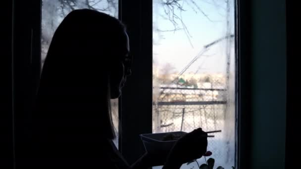 Το κορίτσι στέκεται στο παράθυρο και τρώει τοπ ράμεν. Τρώγοντας στιγμιαία ζυμαρικά — Αρχείο Βίντεο