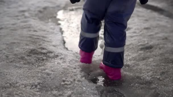 Дитина в рожевих гумових черевиках йде в калюжу з розтопленим снігом. Весняна погода — стокове відео