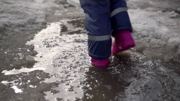 ピンクのゴム製のブーツの子供は溶けた雪の水たまりの中を歩いている。春の天気 ストックフォト