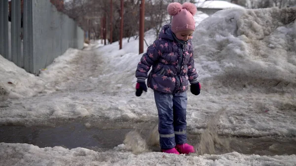 A child in pink rubber boots is jumping in a puddle of melted snow. Spring weather Stock Photo