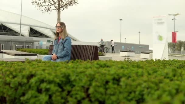 街の庭の緑の花壇の前のベンチに座っているブロンドの女の子 — ストック動画