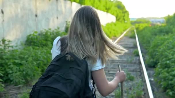 Une fille avec un sac à dos et un bâton dans la main marche le long des traverses. Voyagez sur les voies ferrées. Ancienne voie ferrée — Video