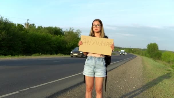 Uma menina com uma mochila fica perto da estrada e segura um sinal com a inscrição onde quer que vá. A pedir boleia. Pare o carro com um sinal com a inscrição — Vídeo de Stock