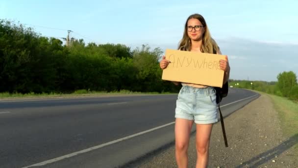 Una chica con una mochila está de pie junto a la carretera y sostiene un cartel con la inscripción dondequiera que vaya. Hacer autostop. Detenga el coche con un cartel con la inscripción — Vídeos de Stock