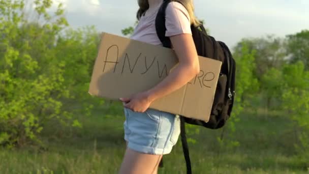 Una chica con una mochila va a pie y sostiene un cartel en la mano con la inscripción ir a cualquier lugar. Hacer autostop. Detenga el coche con un cartel con la inscripción — Vídeos de Stock