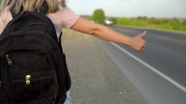 Sırt çantalı bir kız yolun kenarında durur ve baş parmağı havada arabayı yakalar. Otostop yapıyorum. Elinle arabayı durdur.