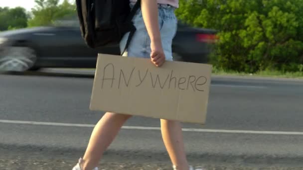 Una chica con una mochila camina por el camino y sostiene un cartel en la mano con las palabras van a cualquier lugar. Hacer autostop. Detenga el coche con un cartel con la inscripción — Vídeos de Stock