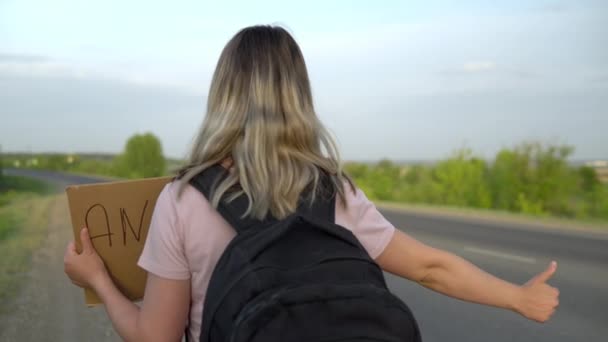 Una chica con una mochila se para junto a la carretera y coge el coche con el pulgar hacia arriba. Hacer autostop. Detén el auto con la mano. — Vídeos de Stock