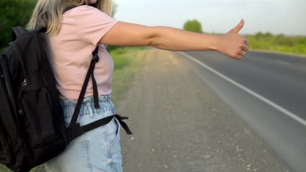 Una chica con una mochila se para junto a la carretera y coge el coche con el pulgar hacia arriba. Hacer autostop. Detén el auto con la mano. — Vídeos de Stock