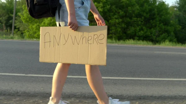 Ein Mädchen mit Rucksack läuft die Straße entlang und hält ein Schild mit der Aufschrift "Überall hin" in der Hand. Per Anhalter. Stoppt das Auto mit einem Schild mit der Aufschrift — Stockfoto