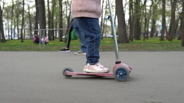 Parkta bir kız asfaltta üç tekerlekli scooter sürüyor.