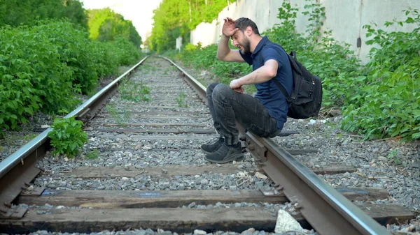 Человек с рюкзаком на спине сел отдохнуть на рельсы. Путешествие по железнодорожным путям. Старый железнодорожный путь — стоковое фото