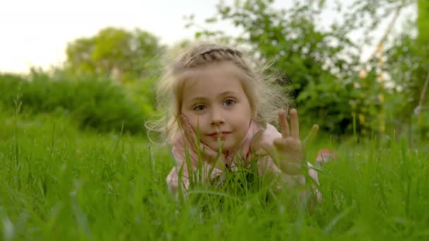 Chica acostada en la hierba agitando sus manos. Niño satisfecho en el verano. 4k — Vídeo de stock