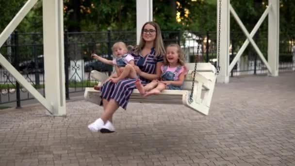 Una donna con due figli sta dondolando su un'altalena. Riposo in famiglia nel parco cittadino. I bambini si siedono con mamma 4k — Video Stock