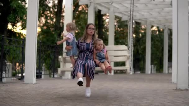 Una donna con due figli sta dondolando su un'altalena. Riposo in famiglia nel parco cittadino. I bambini si siedono con mamma 4k — Video Stock