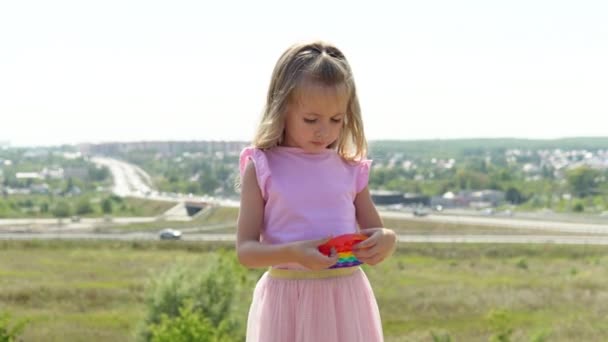 Uma menina de vestido rosa joga popit. A criança é apaixonada pelo anti-stress do arco-íris. Crianças brinquedo de silicone — Vídeo de Stock