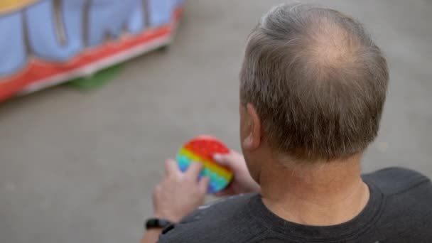 Un uomo di eta 'gioca in un pozzo sullo sfondo di un parco pubblico. La persona è appassionata di antistress arcobaleno. Giocattolo in silicone per bambini — Video Stock