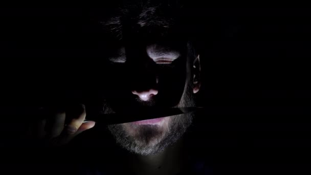 Cechy męskiej, brodatej twarzy, podświetlanej w ciemności. Zbliżenie twarzy mężczyzny w ciemności, który trzyma nóż w zębach — Wideo stockowe